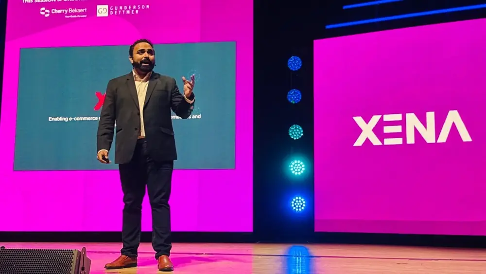Akhil Suresh Nair in a dark blazer pitching on stage between bright pink screens, one showing the XENA logo.