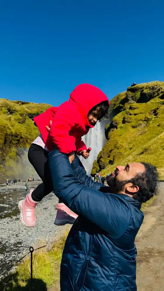 A man lifting a toddler in a red jacket high into the air near a waterfall