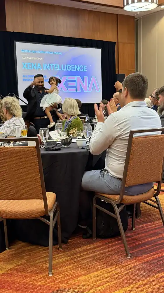 A banquet room with a XENA Intelligence slide on the screen and guests at round tables.